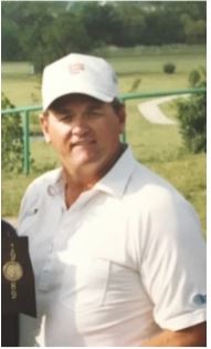 A man in a white golf shirt and cap stands outdoors, smiling, with a golf course visible in the background.